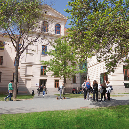 Innenhof der Pädagogischen Hochschule, Menschen sind darauf zu sehen, sie stehen in Gruppen und unterhalten sich, Wiesen und Bäume sind grün, die Fassade weiß